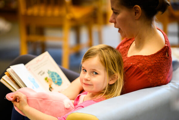 A young girl sitting on a cushioned bench, holding a pink stuffed toy, glances at the camera with a gentle smile. Beside her, an adult reads a children's book aloud. The background shows a cozy library setting with wooden chairs and tables.