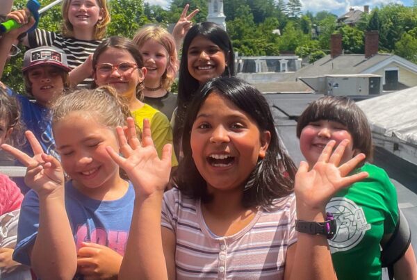 A group of smiling children gathered outdoors on a sunny day, making cheerful gestures like peace signs and waving hands. They are standing close together, with a backdrop of lush green trees and rooftops under a bright blue sky.