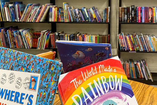 A colorful children's section of the KHL Book Shop is shown. In the foreground, several picture books are displayed on a wooden table, including "Where’s Waldo?" with its signature blue and red patterned cover, and "The World Made a Rainbow," which features an illustrated cover of diverse children playing under a rainbow. In the background, tall black bookshelves are filled with a variety of children's and middle grade books, some arranged upright and others leaning or stacked. The overall scene is vibrant and inviting.